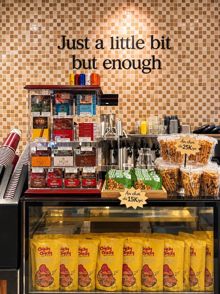 Tủ mica nhiều tầng đựng hộp Toscanello và cigar mini trên quầy BIT Coffee, cạnh snack và đồ uống takeaway tại KDC Chánh Nghĩa